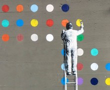 Man painting coloured circles disrupting the plain grey wall