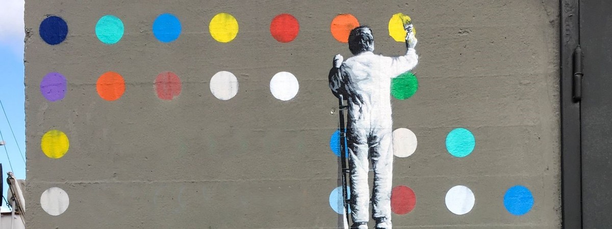 Man painting coloured circles disrupting the plain grey wall