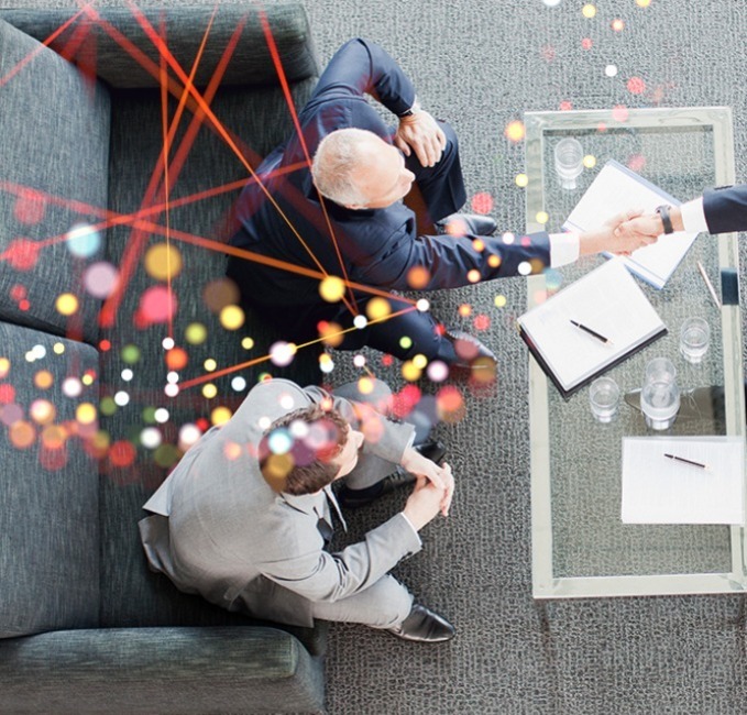 A top-down view of three suited professionals seated around a small table, shaking hands; abstract coloured lines overlay the meeting scene.