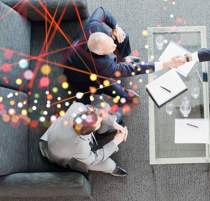  A top-down view of three suited professionals seated around a small table, shaking hands; abstract coloured lines overlay the meeting scene.