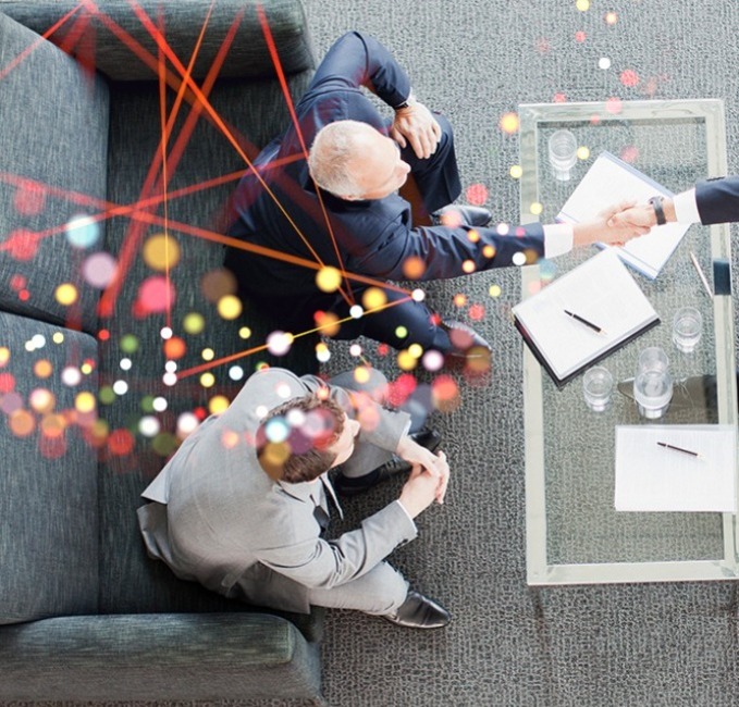 A top-down view of three suited professionals seated around a small table, shaking hands; abstract coloured lines overlay the meeting scene.